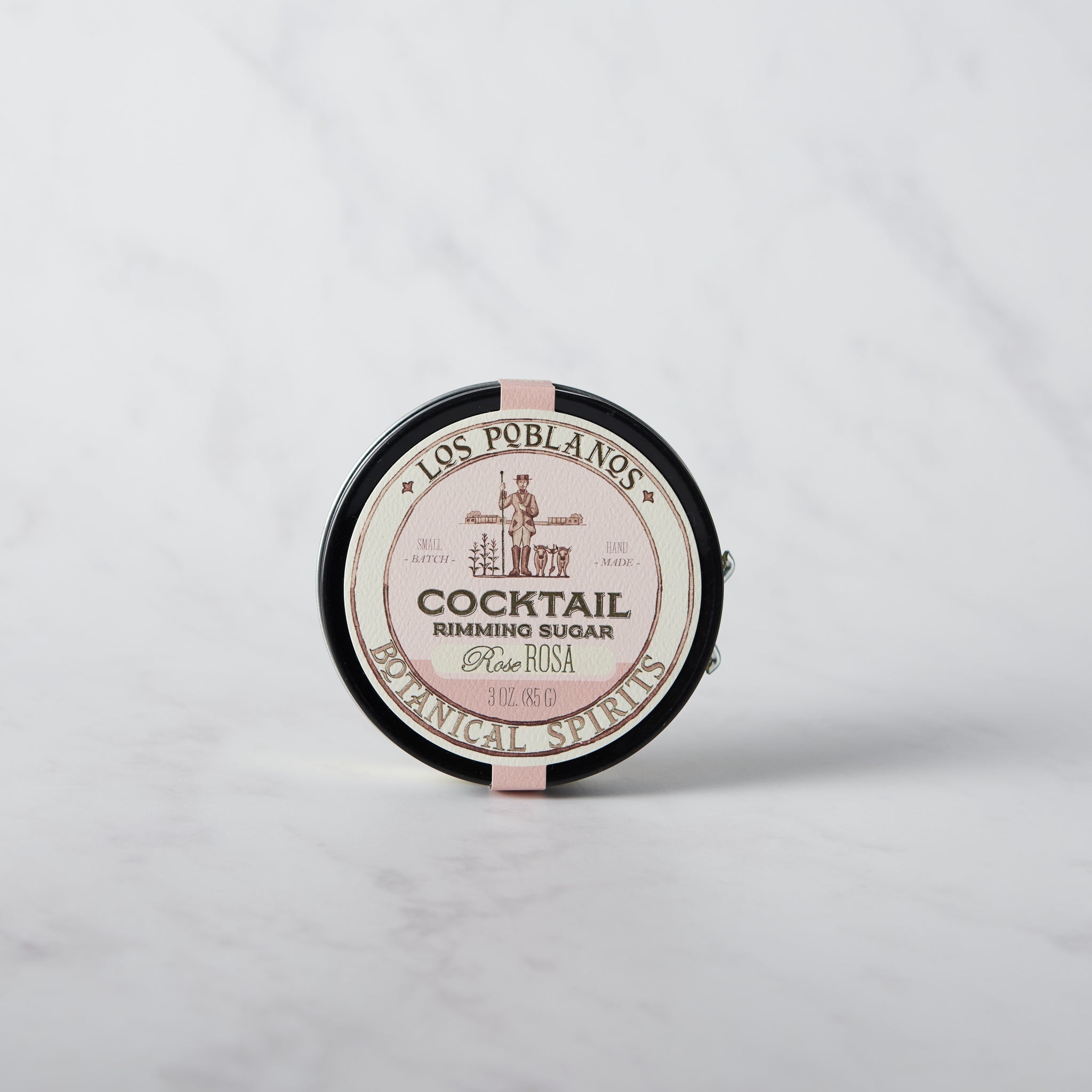 A round tin container of Los Poblanos Cocktail Rimming Sugar with rose flavor, placed on a marble surface.
