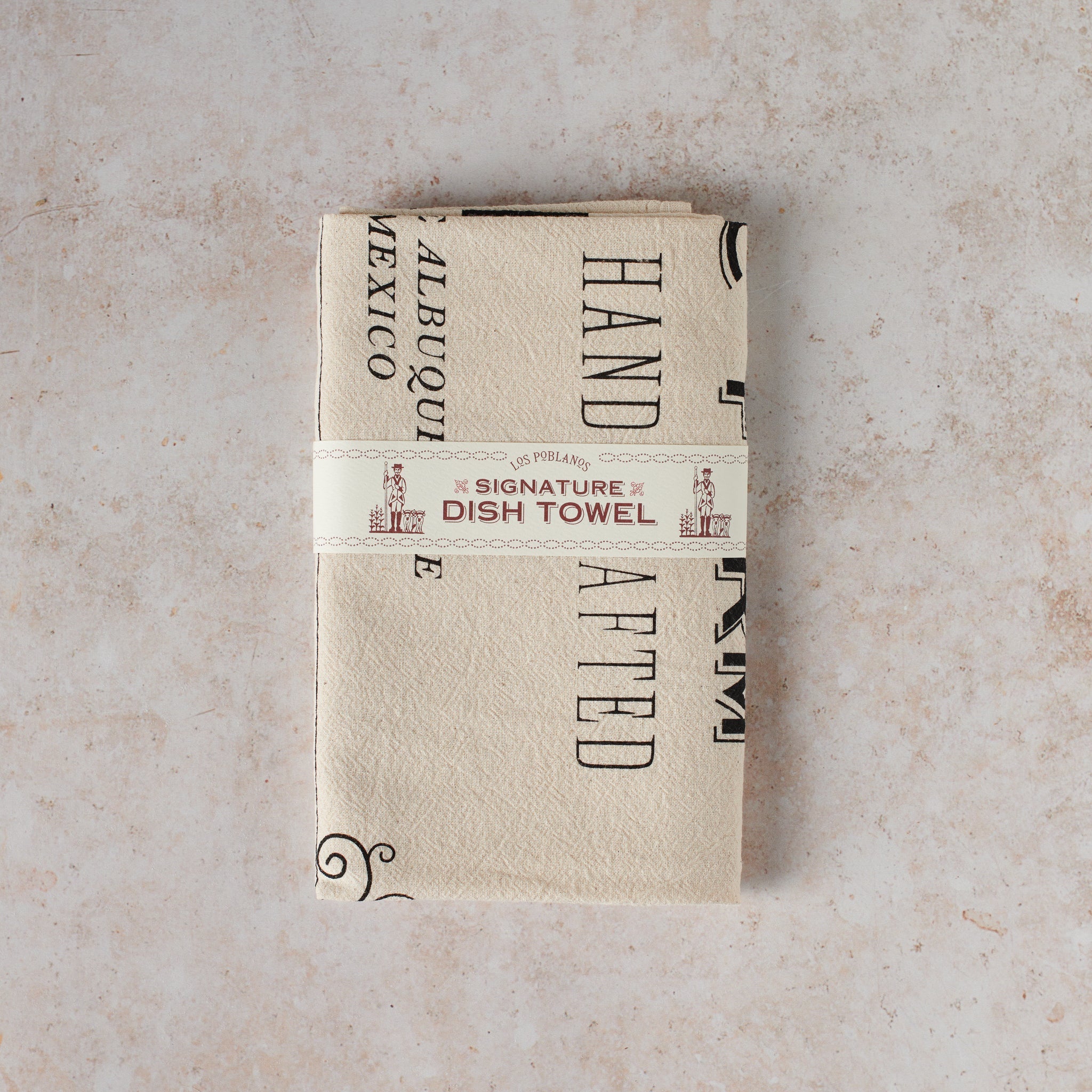 Folded beige cotton dish towel with text and design elements, handcrafted in New Mexico, wrapped and ready for gifting.
