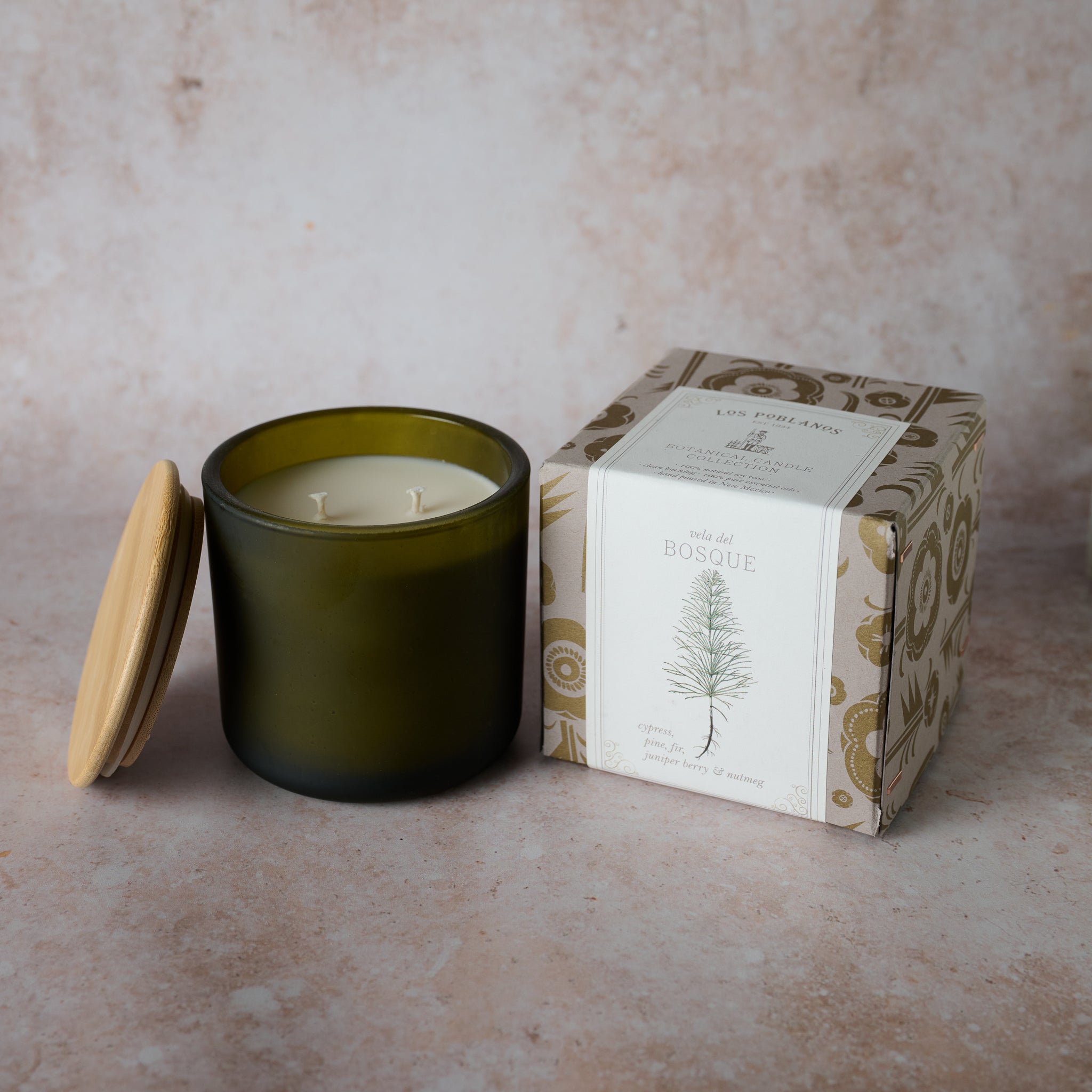 A green frosted glass candle vessel with a wooden lid next to a box that has an illustration of a tree. The candle is unlit and appears to have a woodsy scent, possibly pine and fir.