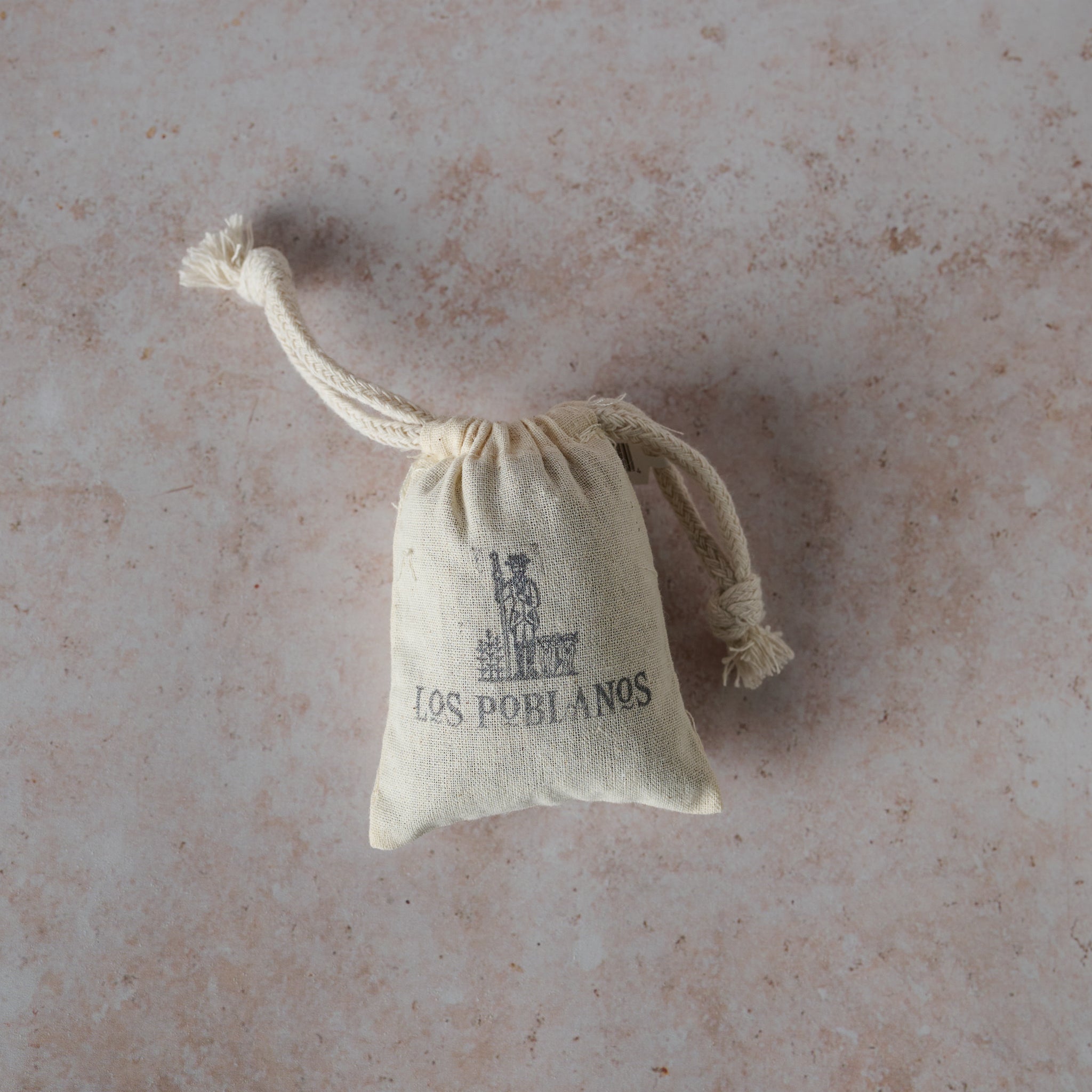A small hand-stamped muslin bag with drawstrings, printed with 'LOS POBLANOS', filled with dried lavender buds.