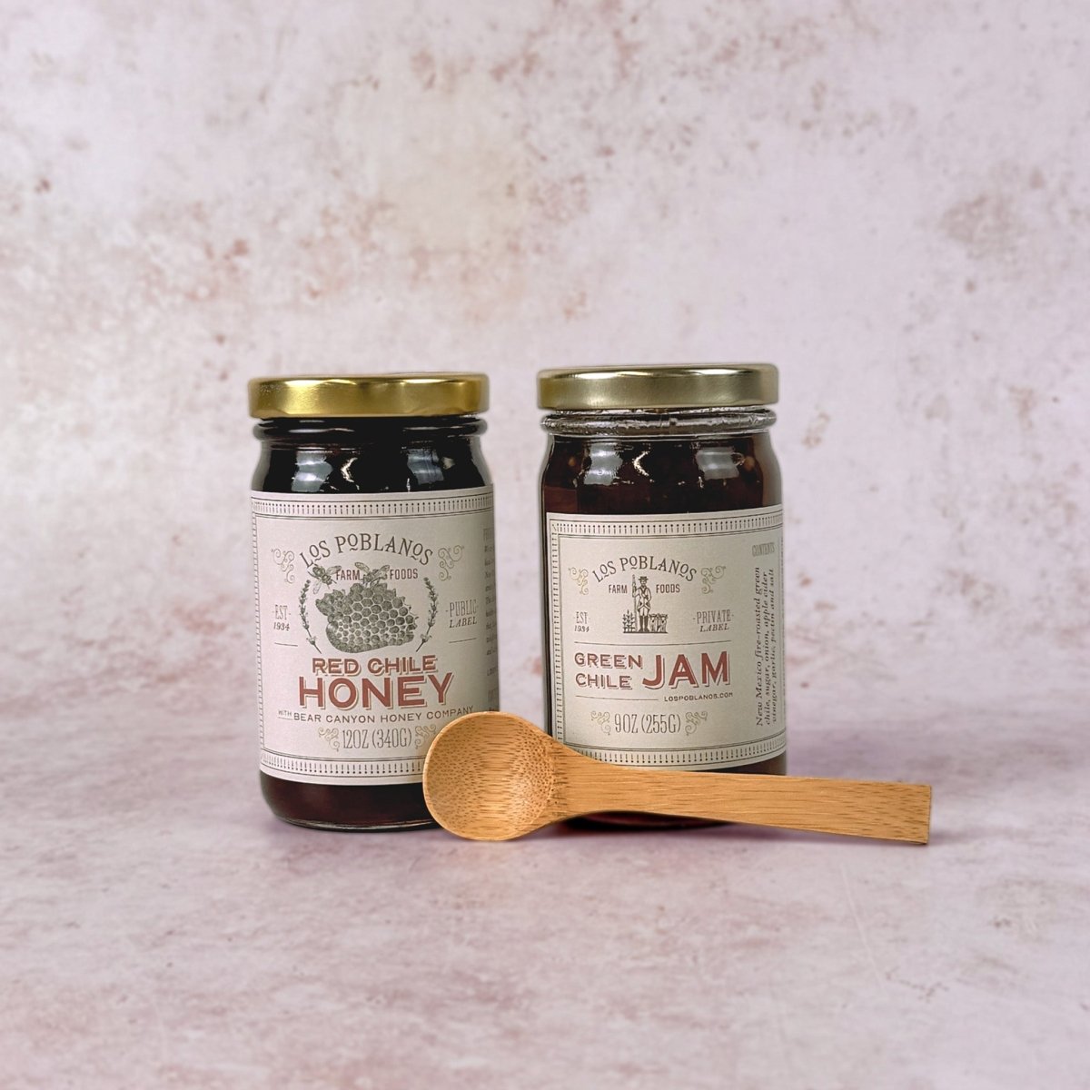 Two jars of Los Poblanos red and green chile honey and jam with a wooden spoon on a textured surface.