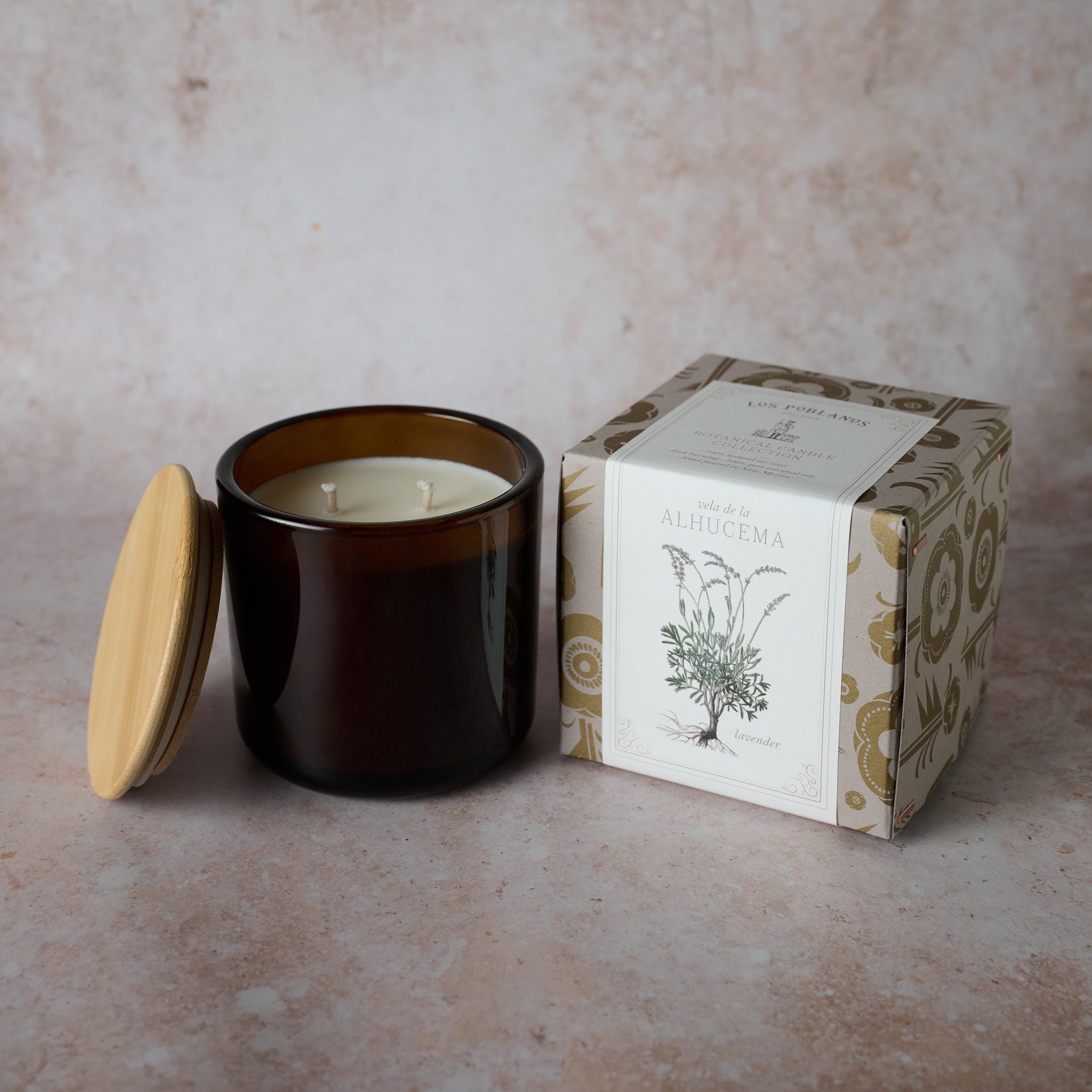 A soy wax candle in an amber glass jar with a wooden lid, next to a box with a lavender illustration. The candle is white and the box is adorned with a floral pattern.
