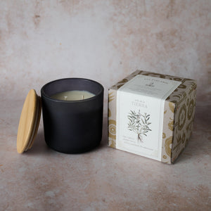 A black soy wax candle with a wooden lid next to its packaging box, which has a botanical design. The candle is unlit and appears to be in a frosted glass container.